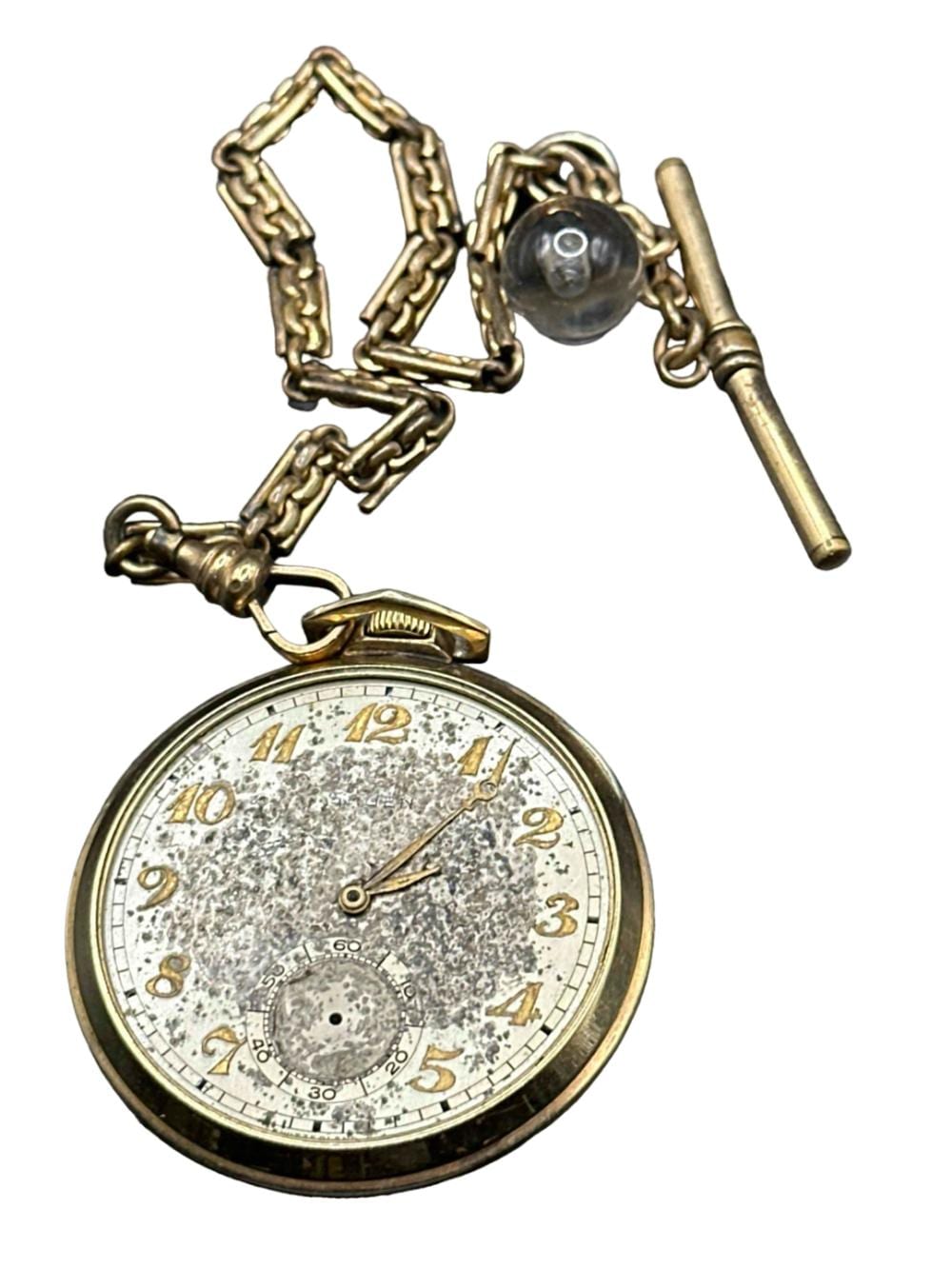 Open-face vintage pocket watch with a chain and key wind stem, showing a speckled white dial with gold hour numerals and black minute track.