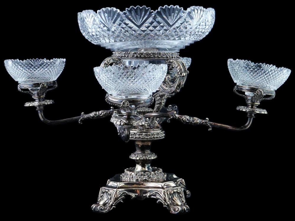 Silver-colored ornate pedestal stand with a faceted glass crystal bowl and two matching smaller glass cups on either side, on a dark background.