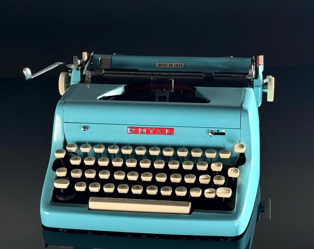 A teal vintage typewriter with cream keys and a paper tray, shown front-on against a dark background.