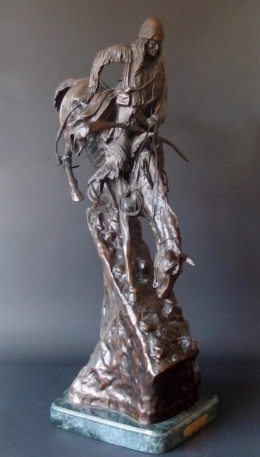 Bronze sculpture of a cowboy-style figure seated or leaning forward, holding a rope with a hat on his head, with a lasso in front.