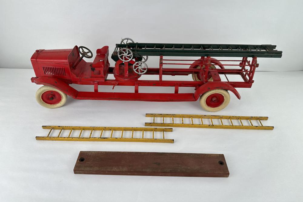 Red toy fire ladder truck with white side panel, wheels, and extended ladder; separate ladder sections and brown base plate laid out below.