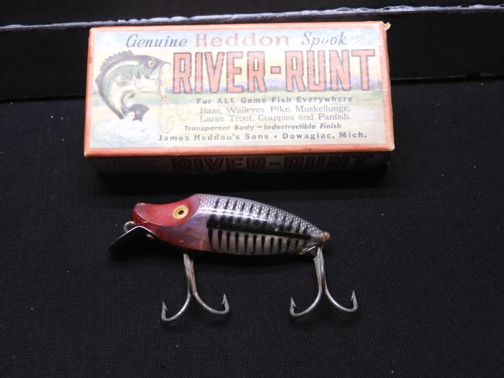 A fishing lure with a red, white, and black painted body sits on a dark surface in front of its boxed label reading “Genuine Heddon Spook River-Runt.”