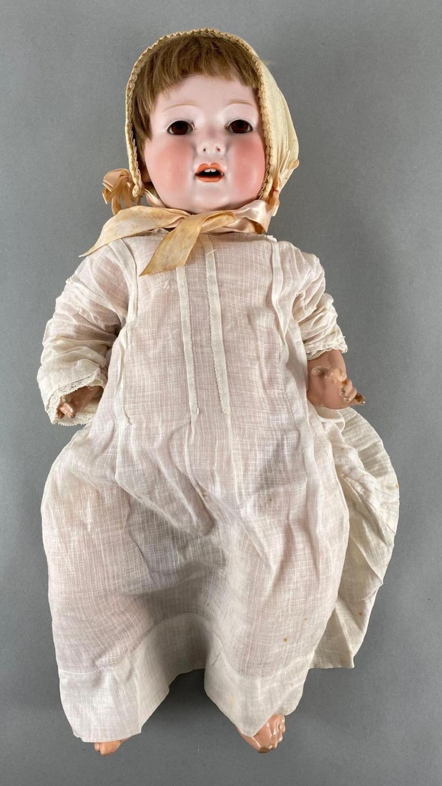 Porcelain baby doll with an open mouth, brown molded hair, a beige hood and bow, and a white patterned dress against a gray background.