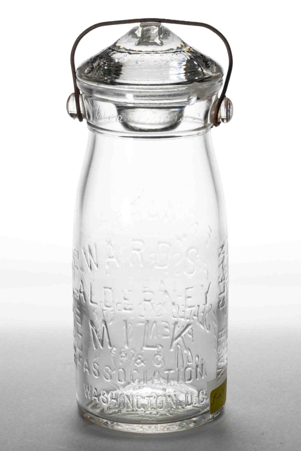 Clear glass milk bottle or preserving jar with bail handle and metal clamp lid, embossed with text and decorative pattern.