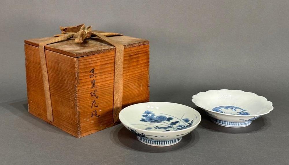 Two blue-and-white porcelain dishes with scalloped rims, shown on a gray background next to a wooden box tied with a cloth ribbon.