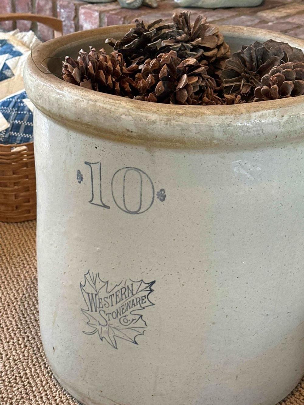 Large white stoneware crock with a painted “10” and pinecone decorations, filled with pinecones.