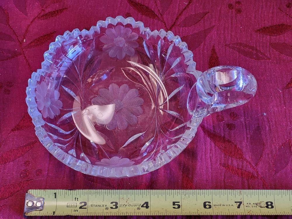 Round clear glass candy dish with scalloped rim, cut floral and fan designs, and a heart-shaped clasp-like form handle on the side.