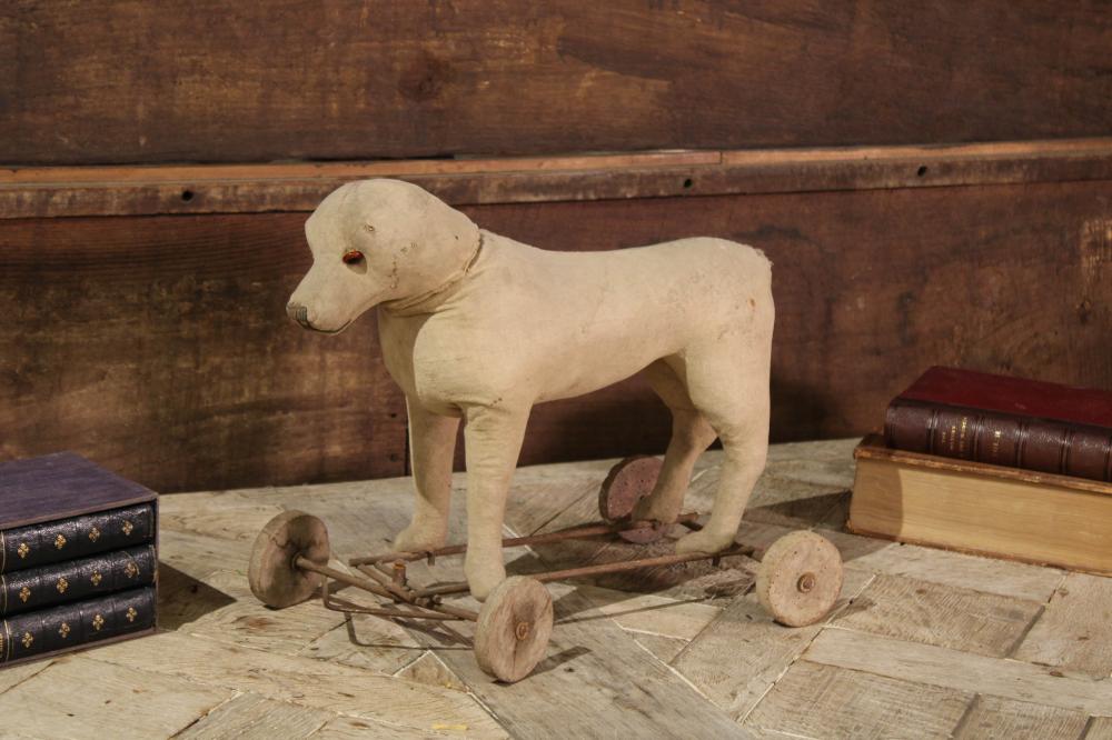 White wooden toy dog with painted red eyes standing on wheels on a tabletop in front of books.