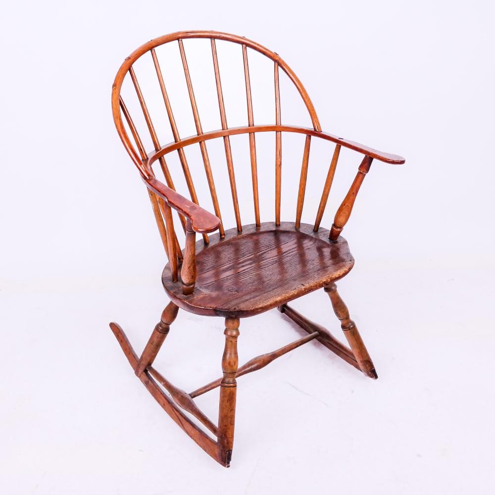 Wooden rocking chair with curved spindle back and round seat, shown at an angle against a white background.