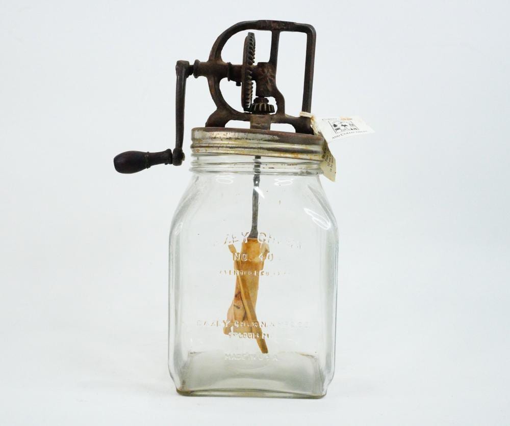 Glass jar butter churn with an ornate metal crank and plunger, shown upright against a light background.