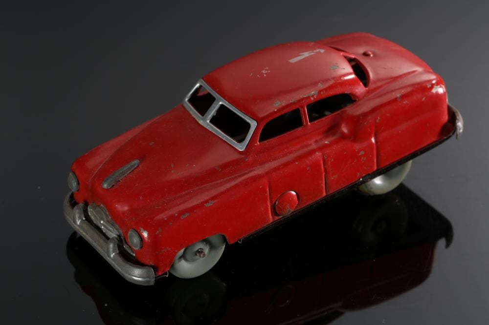 Red vintage tin wind-up toy car with gray wheels and a open-top window detail, shown on a dark reflective surface.