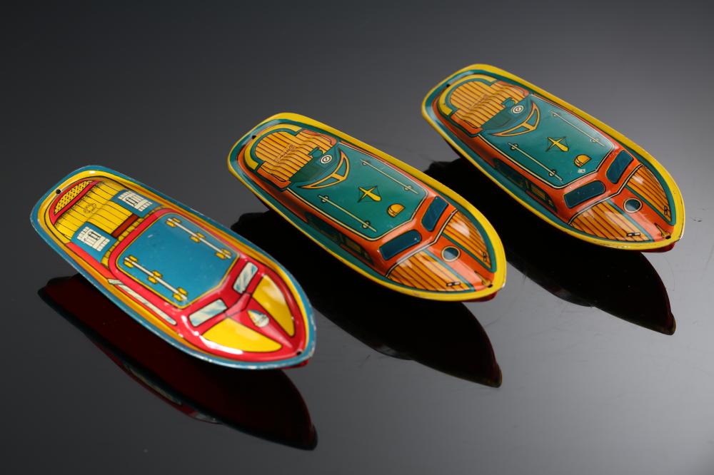 Three colorful tin lithograph toy boats with teal panels, yellow striped sides, and red accents, photographed on a dark background.