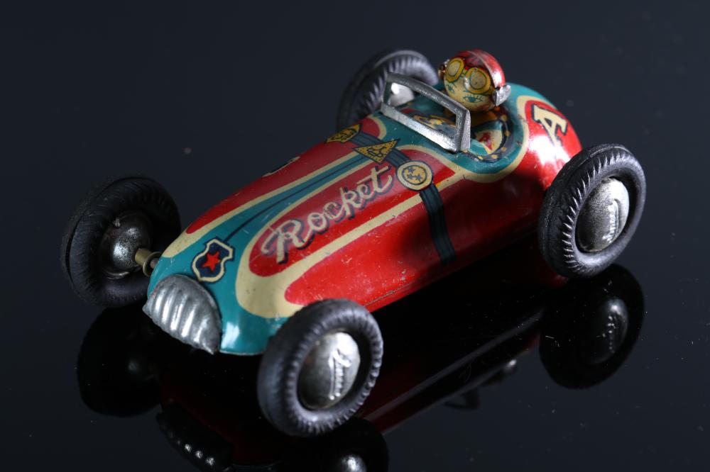 Colorful tin friction rocket-racer toy car with winged graphics, black tires, and a small character cockpit, photographed on a dark background.