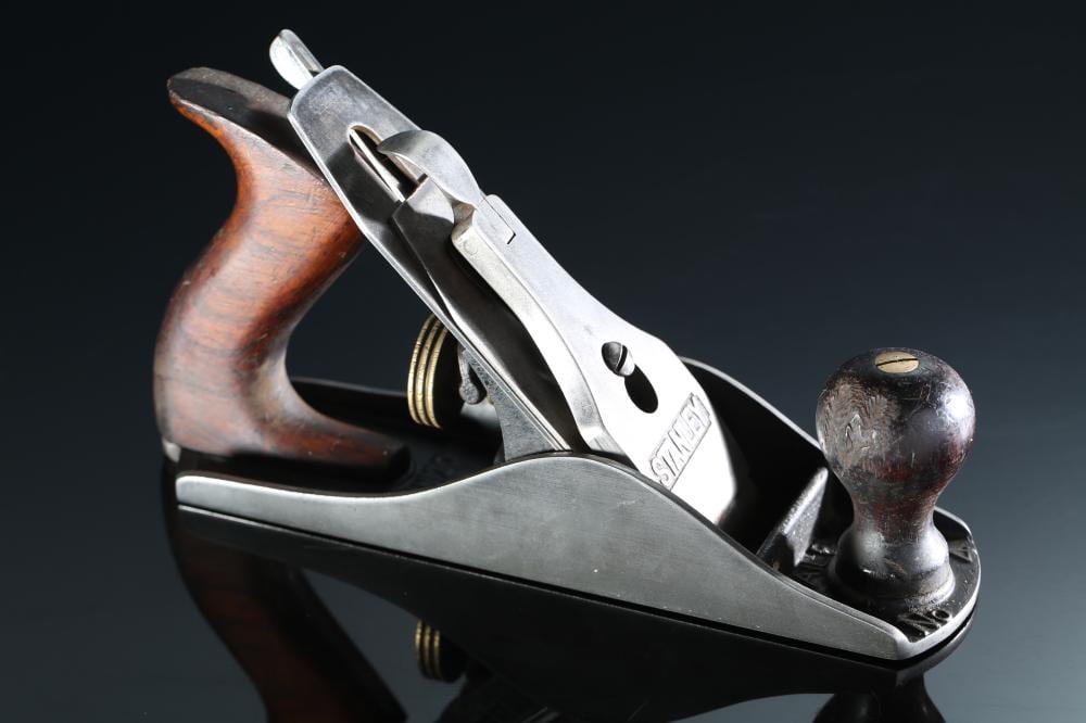 Vintage hand wood plane with metal body, wooden handle, and side knob against a dark background.