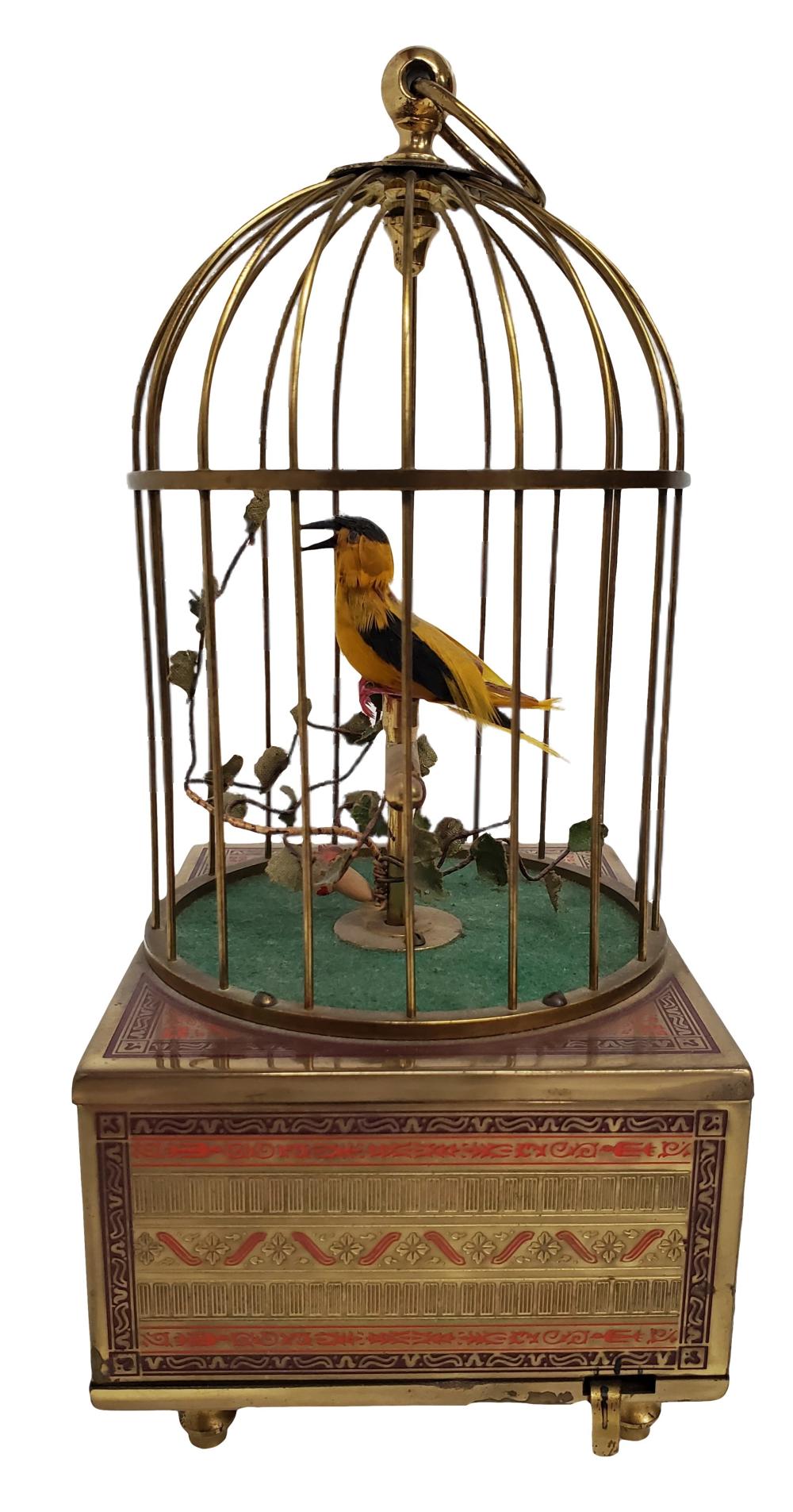 Antique brass birdcage music box with a yellow singing bird inside, surrounded by arched bars and a green base, photographed on a white background.