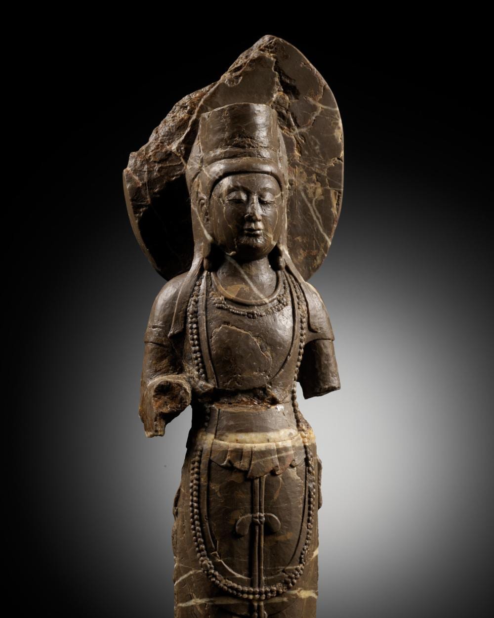 Stone sculpture of a seated standing Buddhist deity with a halo behind the head, wearing layered robes and jewelry.