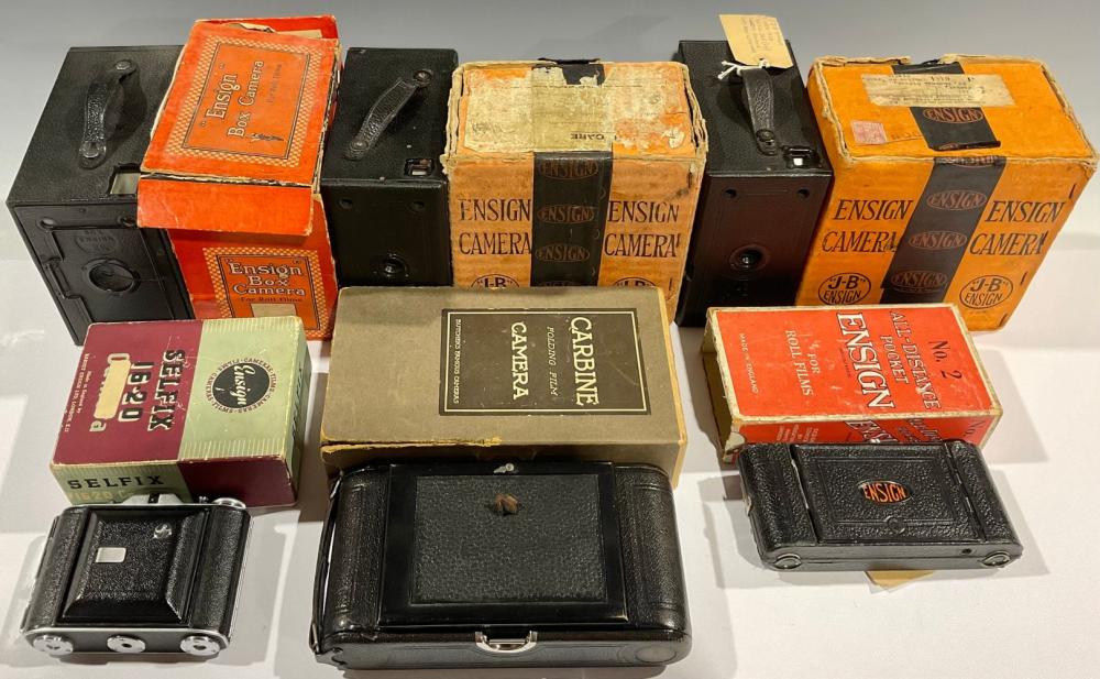 Group photo of several vintage box cameras and their colorful retail boxes laid out on a white surface, including Ensign-branded cameras.