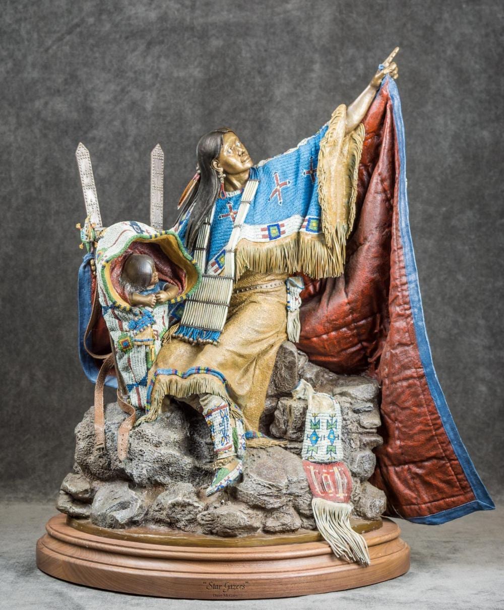 Bronze-style figure sculpture of a native woman standing on a rocky base, holding up one arm while wrapped in patterned fabric and a red-blue cloak.