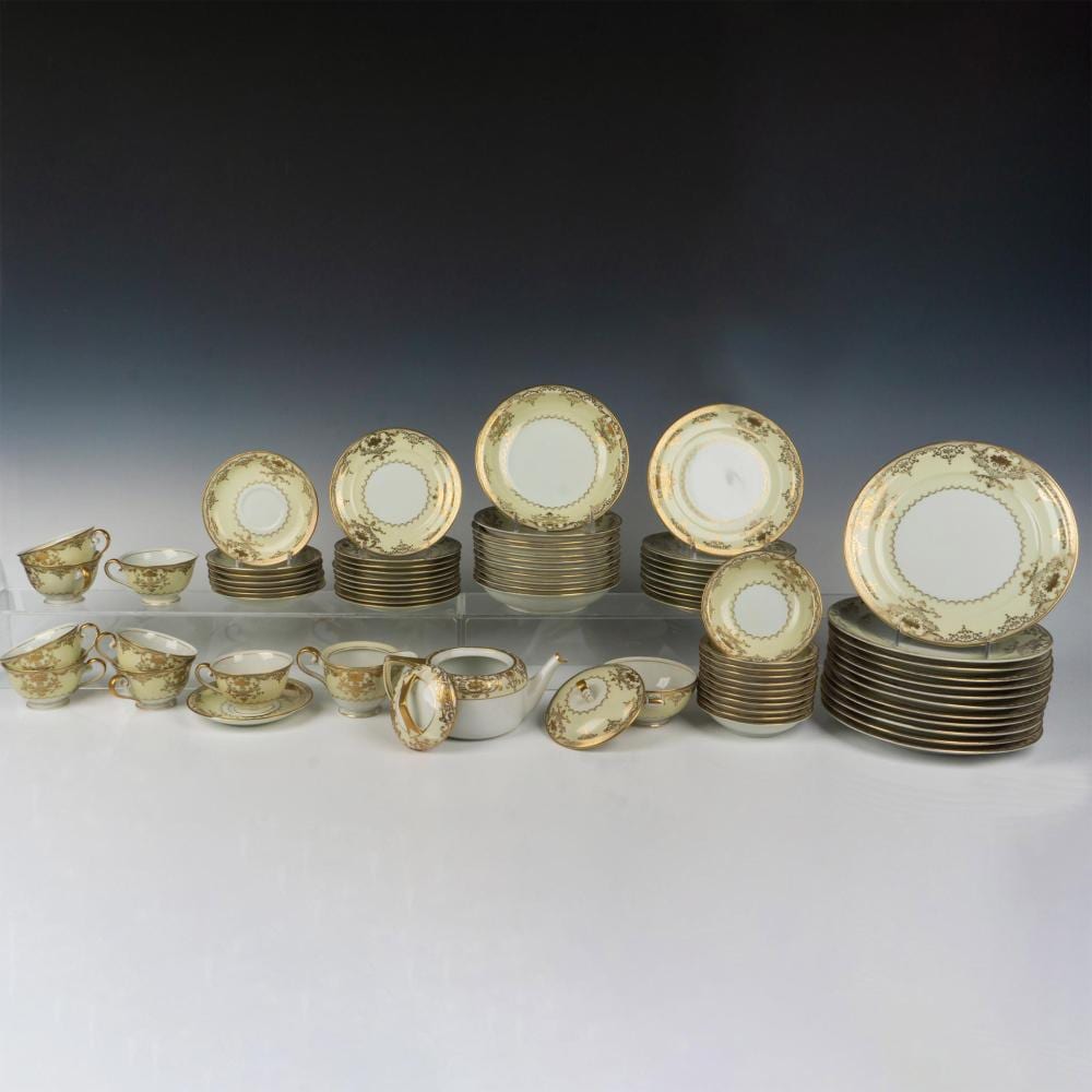 Set of gold-trimmed white porcelain dinnerware pieces, including stacked plates, cups, saucers, and a cream pitcher on a light tabletop.
