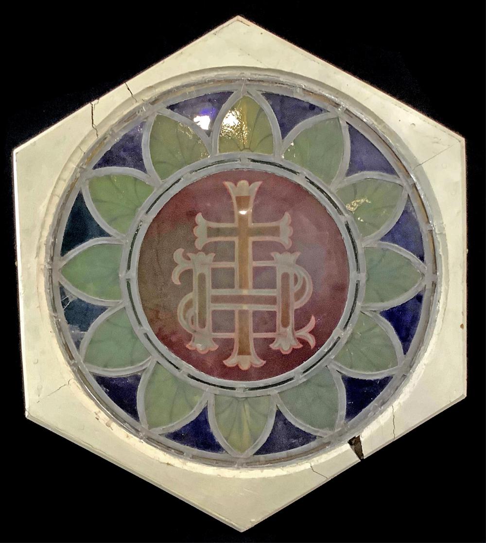 Hexagonal stained glass window panel with blue and green quatrefoil shapes and a central red roundel featuring a white monogram cross.