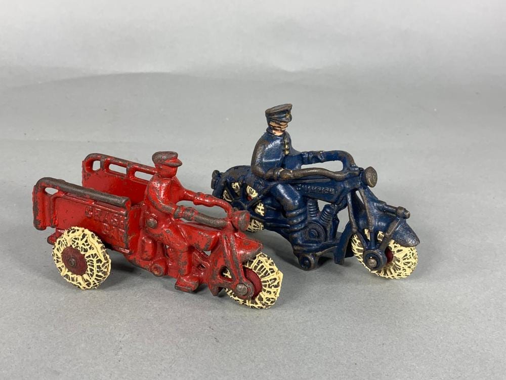 Two small cast-iron toy motorcycles on a gray background, one red and one blue with a rider figure and gold-toned wheels.