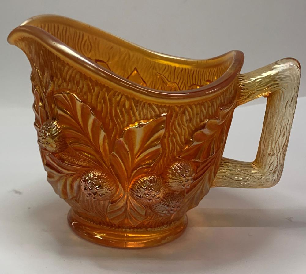 Amber glass creamer with carved floral and leaf pattern, clear handle, and scalloped rim.