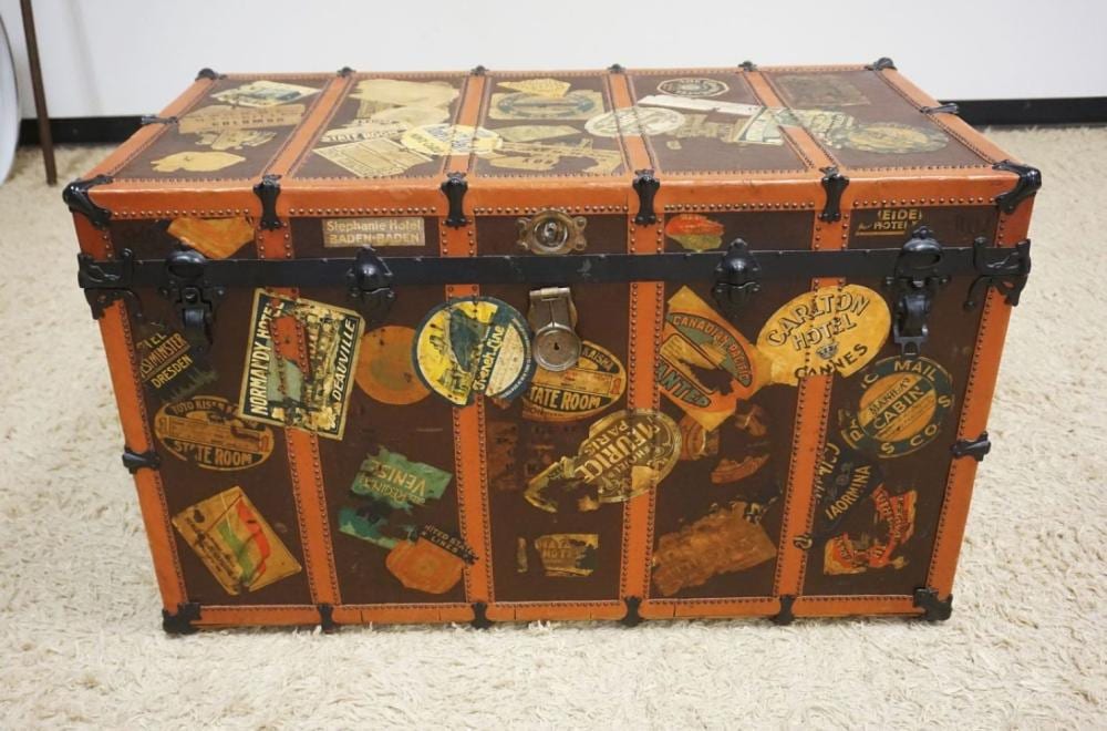 Orange vintage travel trunk with metal corner hardware, studs, and patchwork luggage stickers on the front and lid.