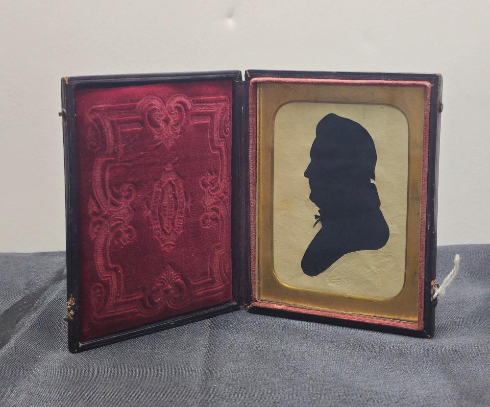 Open framed silhouette portrait showing a black profile bust on a light background, inside a decorative dark frame with gold edging.