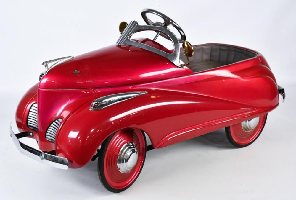 Red pedal car with chrome hood ornament, curved fenders, spoked wheels, and an open cockpit with steering wheel.