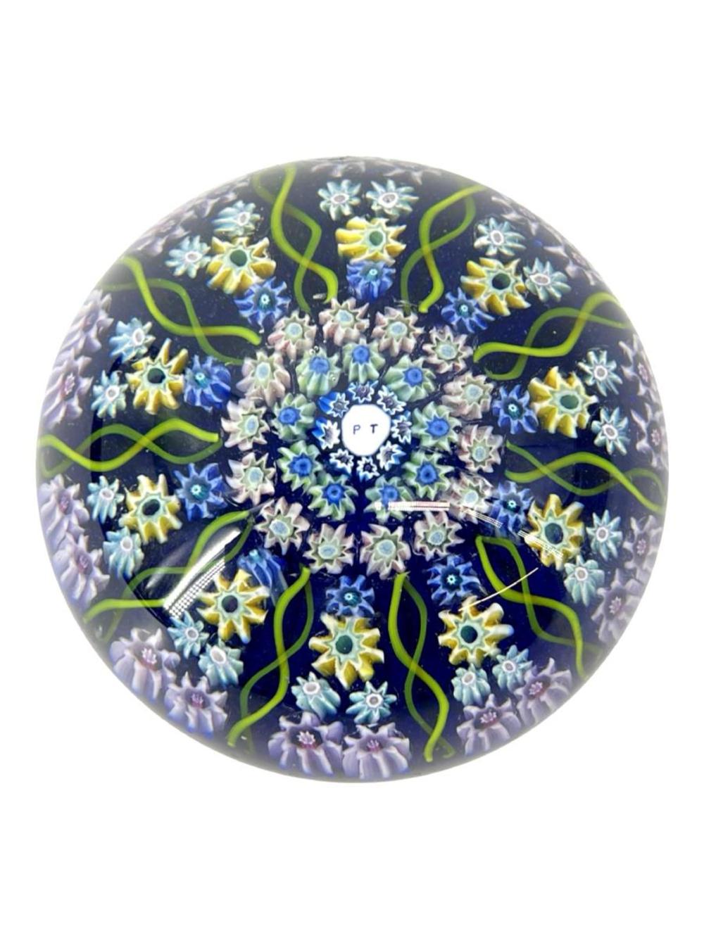 Round blue glass paperweight filled with colorful starburst and flower-like millefiori patterns and green curving lines.