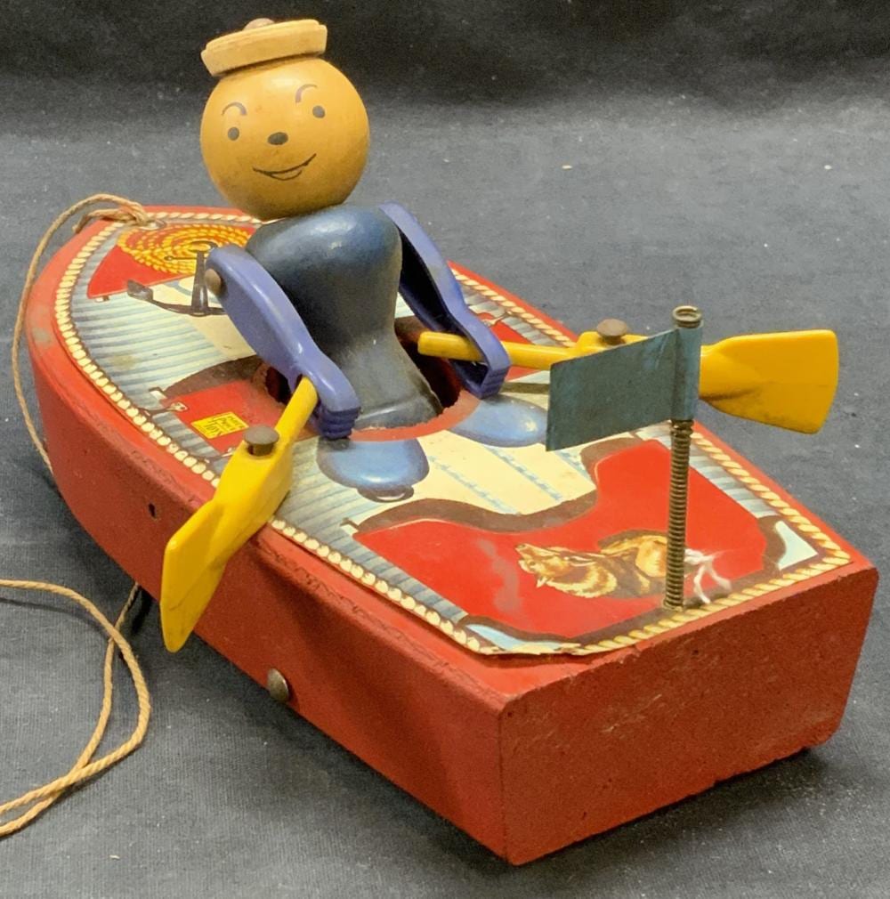 Vintage toy pull boat with a smiling yellow-headed driver, blue pants, and colorful sailboat-like parts on a red and blue printed boat base.