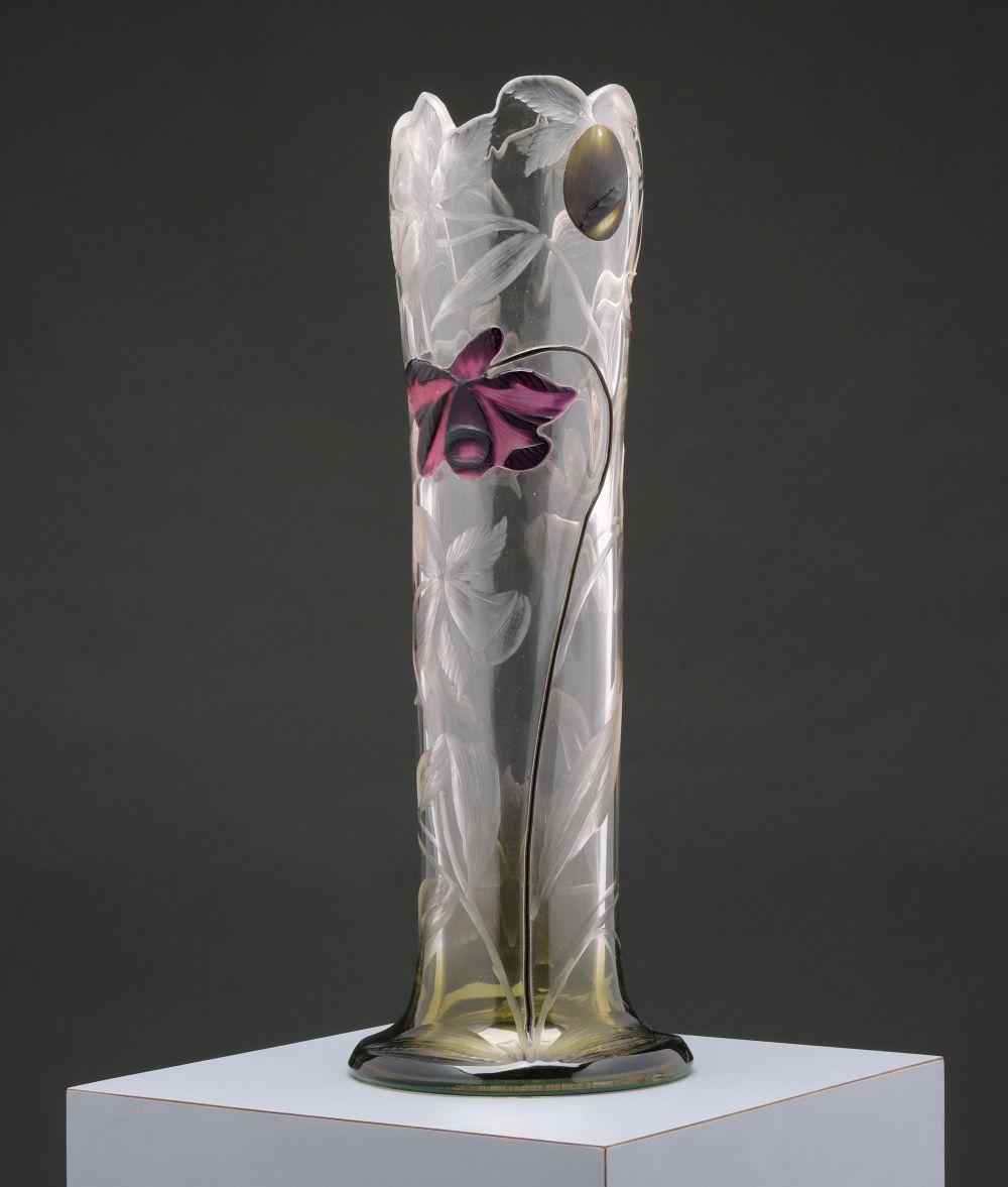 Tall clear glass vase with raised floral and leaf relief, a purple flower accent, and a gold-toned oval inset at the rim.