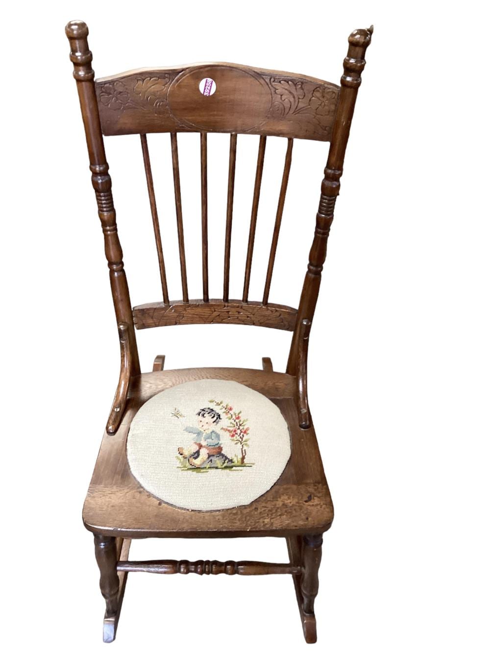 Wooden rocking chair with turned posts and a round needlepoint-style seat cushion featuring a small child and flowers.