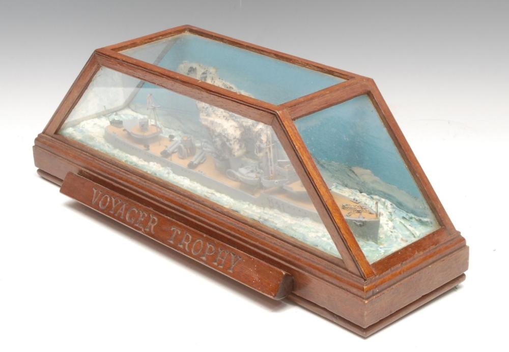 Wooden display case containing a miniature model ship on a glass top, with printed ocean-and-harbor scene panels and the name “VOYAGER TROPHY.”