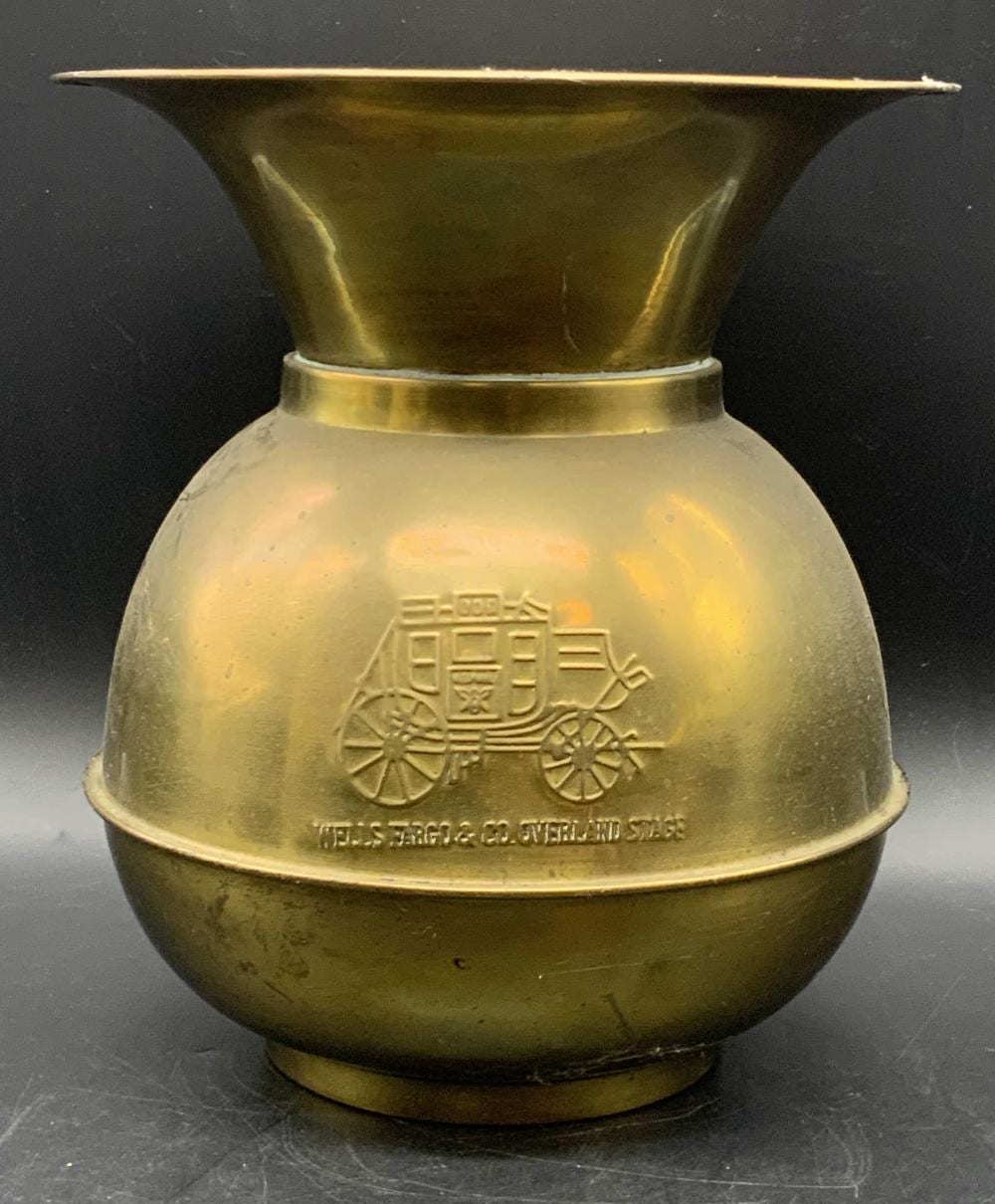 Brass spittoon with a round body and flared rim, showing a raised logo of a wagon and the word “WELLS FARGO”.