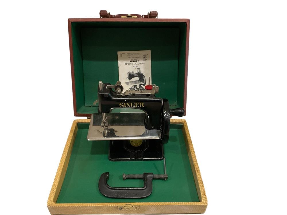 Singer tabletop sewing machine with metal body and hand crank accessories inside a brown open case lined with green fabric.