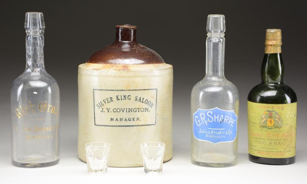 Group of four beverage bottles and a ceramic whiskey jug on a dark background, including a jug labeled "Silver King Saloon" and "J.Y. Covington".