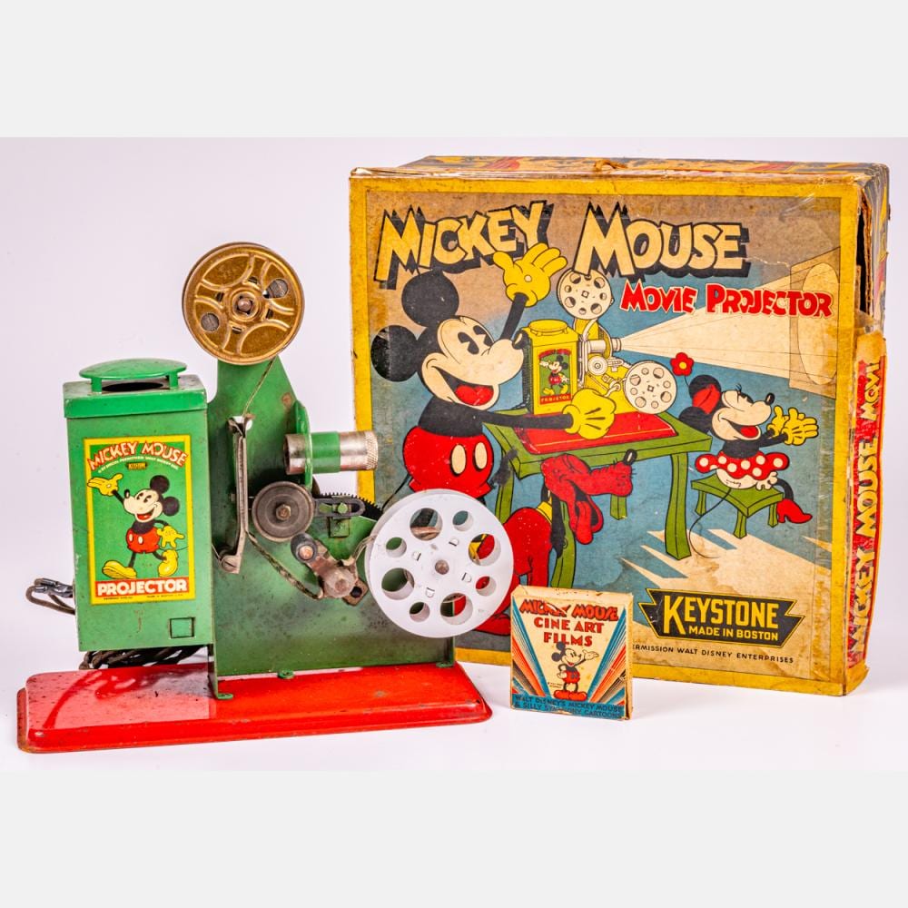 Green vintage toy movie projector with Mickey Mouse artwork, film reel, and round viewing lens, next to its cardboard box.