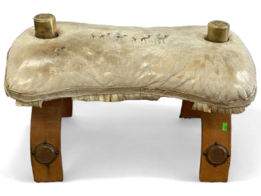 Vintage camel saddle-shaped stool with a light fabric seat and brass-colored corner mounts on a brown wooden base.