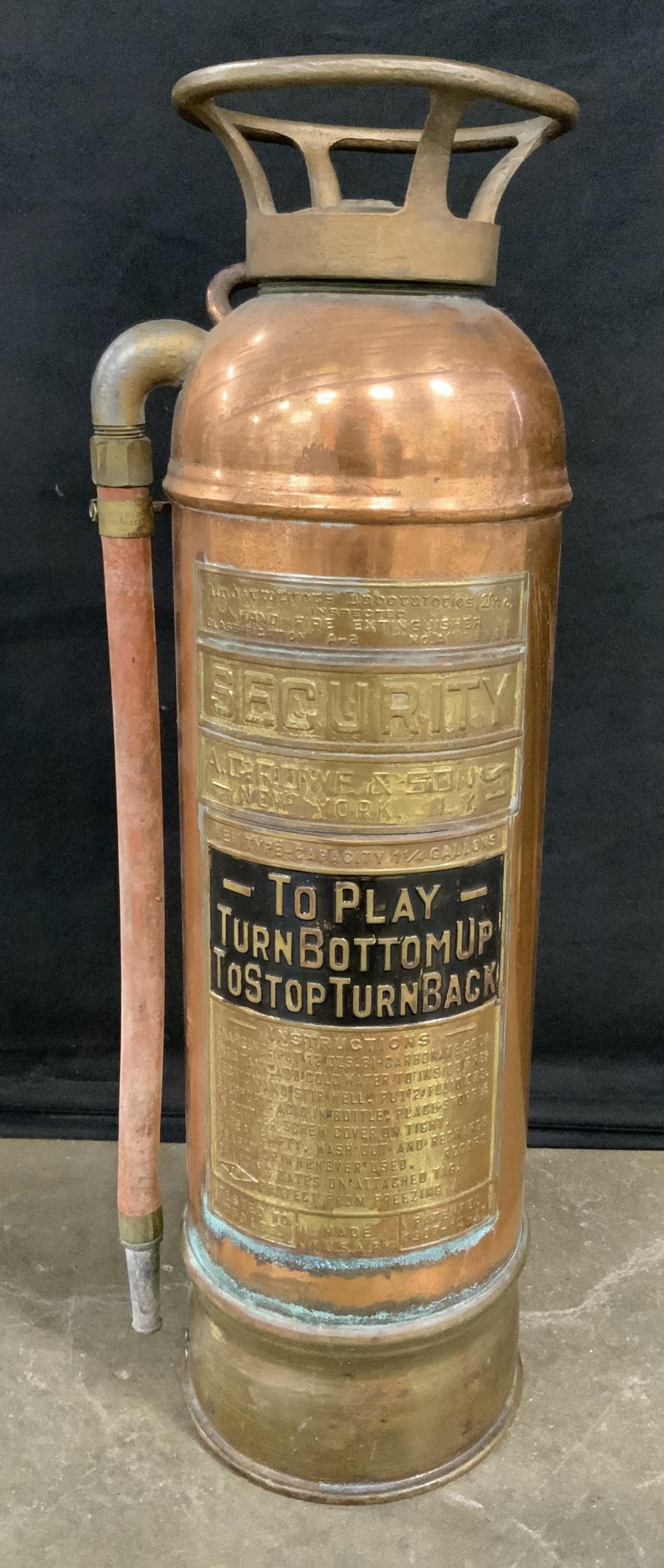 Vintage copper-colored fire extinguisher with metal handle and nozzle, shown from the front against a dark background.