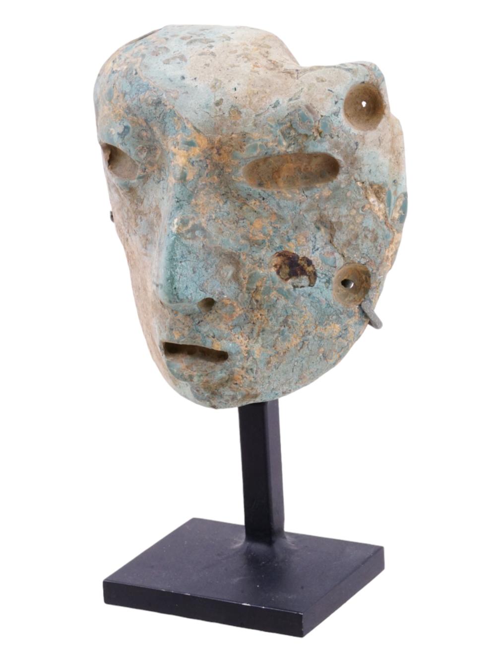 Carved stone face mask on a stand, with turquoise-blue and tan mottled surface, eye opening and small holes for attachment.