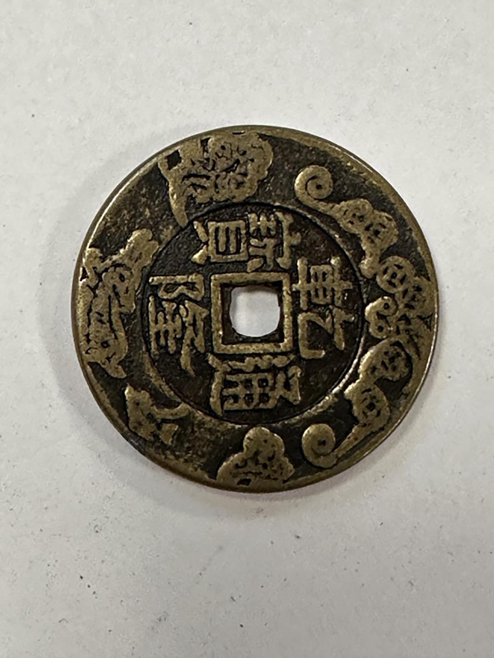 Close-up of an antique-style coin with a square emblem in the center surrounded by decorative symbols and Chinese-style characters, photographed on a white/gray