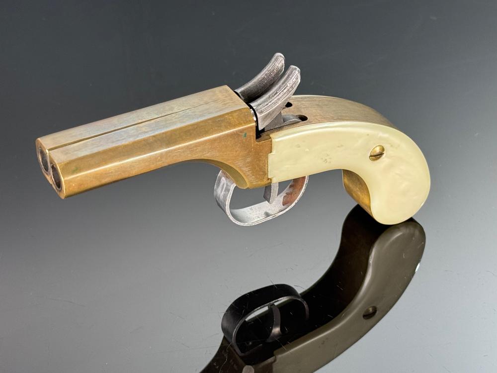 Side view of a small double-barrel percussion pistol derringer with a beige grip and silver metal frame on a reflective display stand.