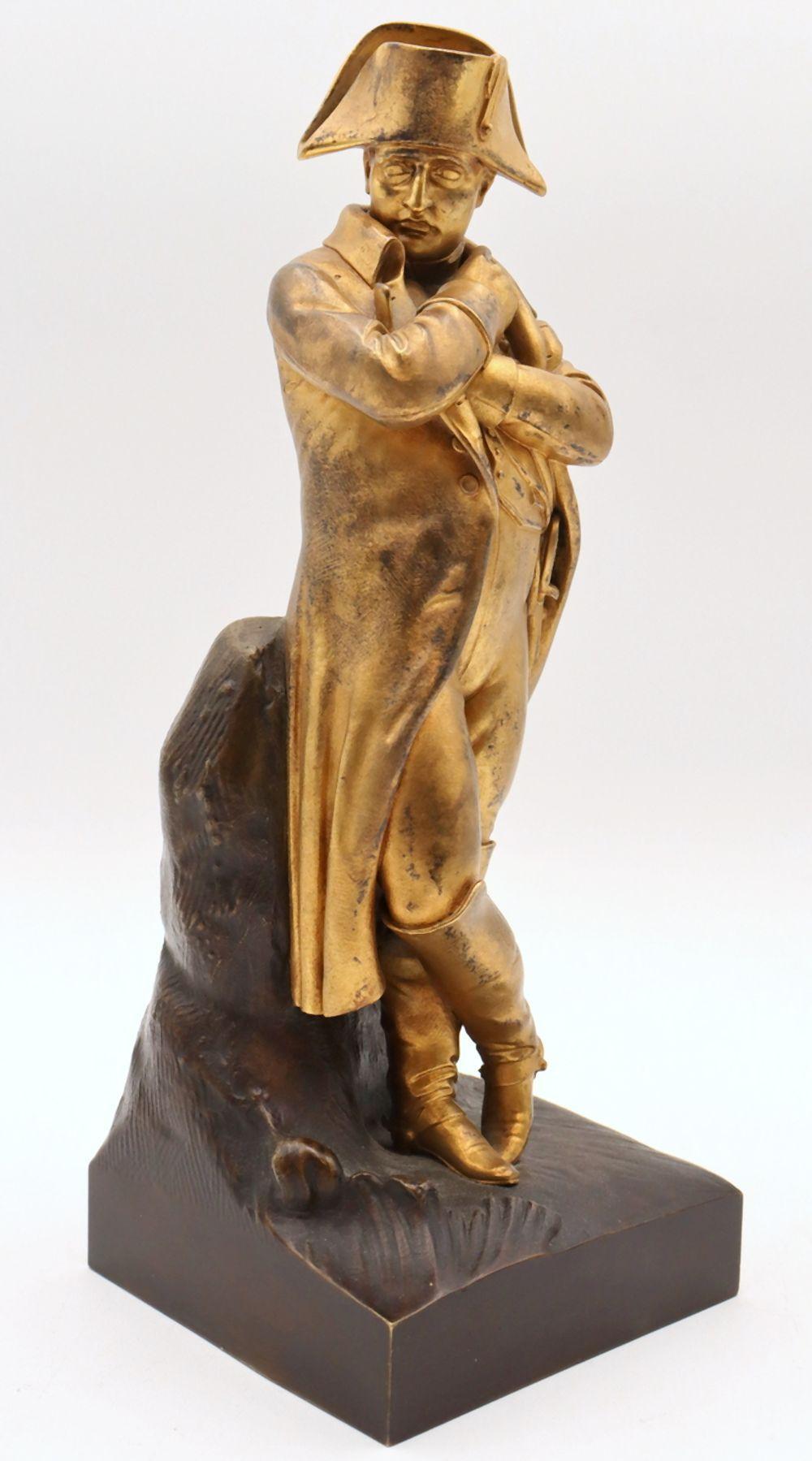 Bronze sculpture of a seated man in a long coat holding an arm across his chest and a hat held over his head on a dark base.