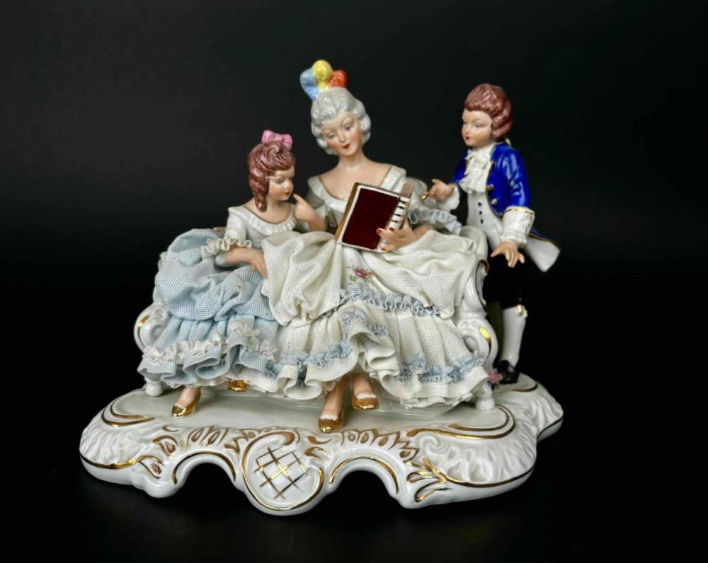 Porcelain figurine group of three children in lace dresses, one reading a red book, on an ornate oval base with painted gold details.