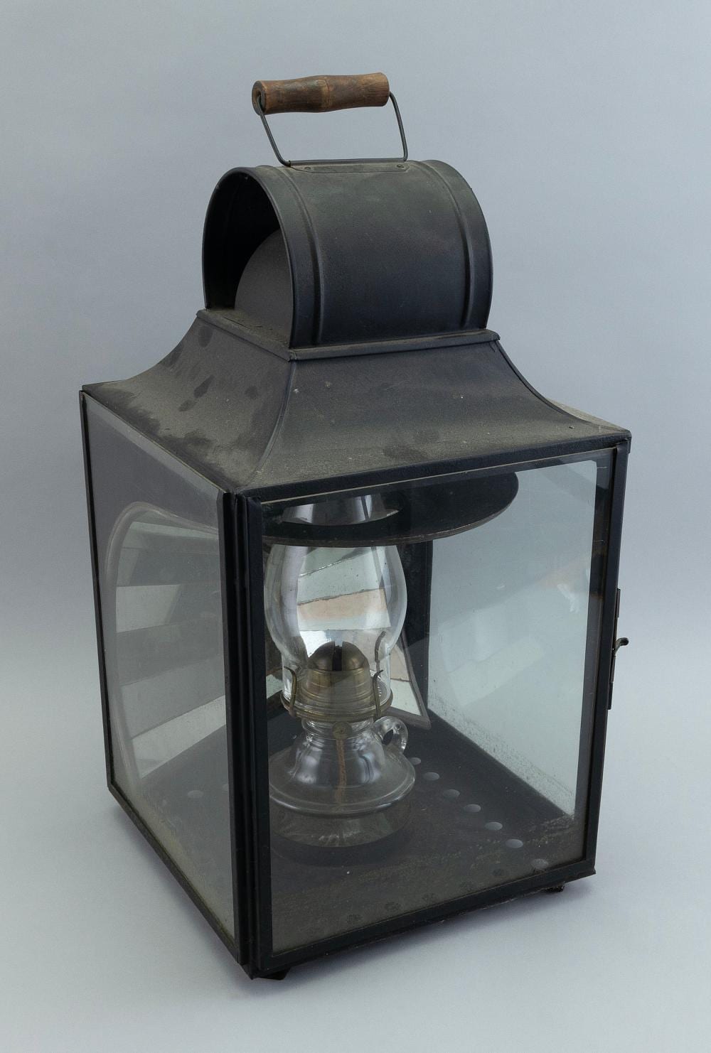 Dark metal barn lantern with glass panels and a clear oil lamp inside, topped with a brass handle and chimney.