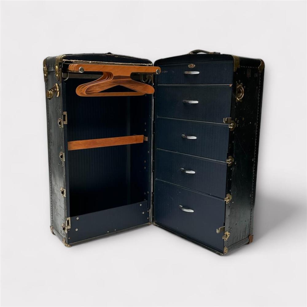 Vintage blue travel trunk shown open, revealing an interior with drawers and a wooden hanger rack, brass corner fittings, and a clasped lid.