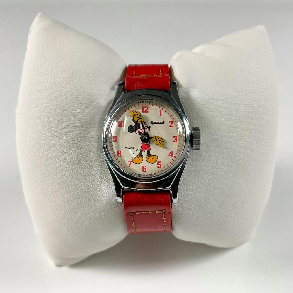 A Mickey Mouse wristwatch with a round white dial and red wristband, shown on a white display cushion.