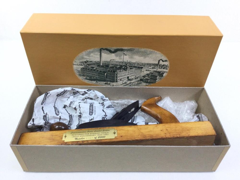 Wood hand plane with metal blade and handles in an open cardboard box with packing paper and a label.