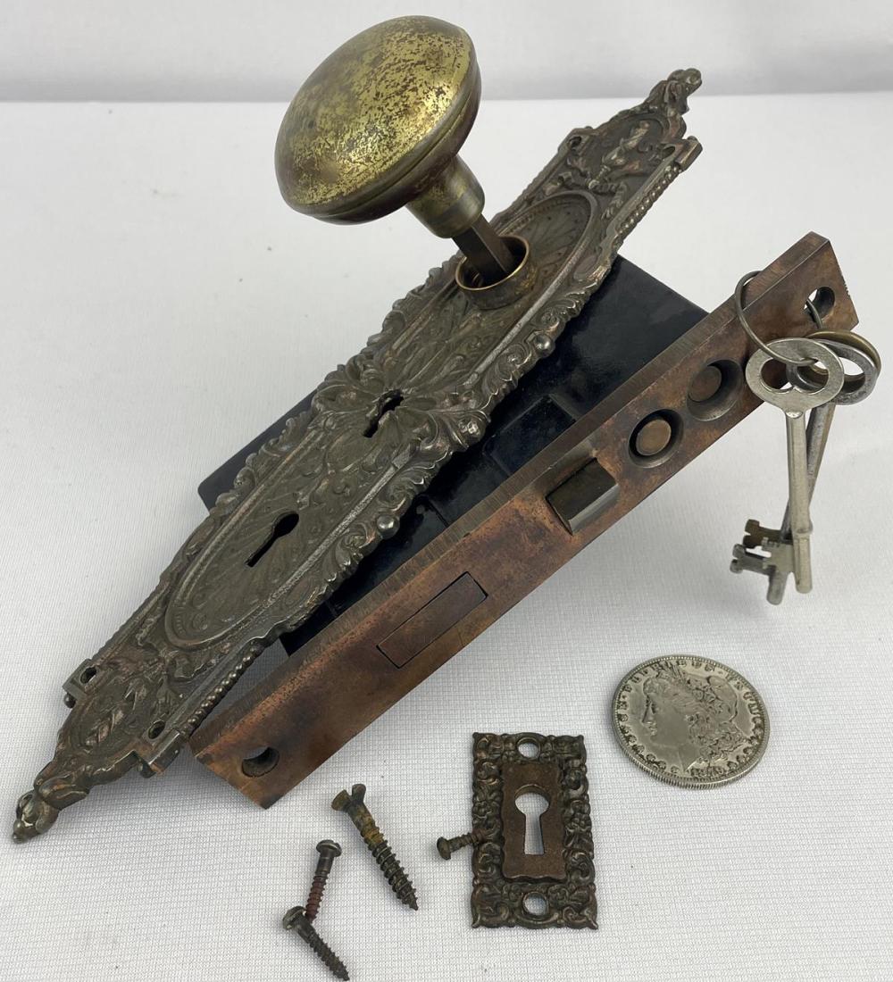 Antique-looking metal mortise door lock set with decorative door plate, doorknob, keyhole cover, and keys on a white background.
